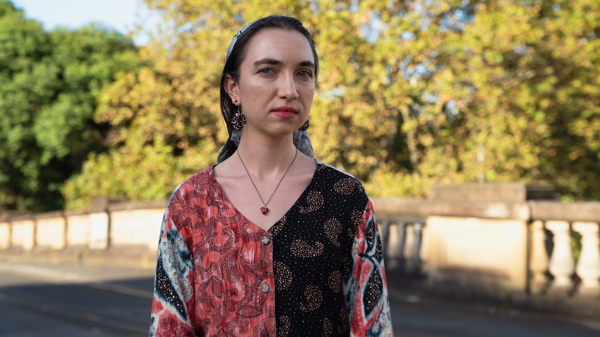 Young woman wearing patterned blouse, with hair pulled back by loose headscarf, standing on bridge with trees in background.