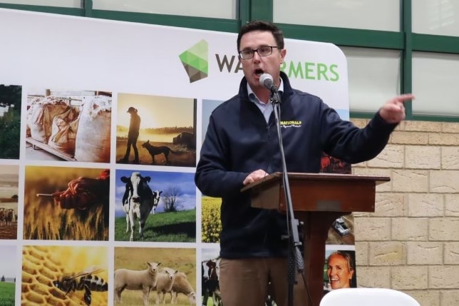 a man in a casual jacket  speaks at a lecturn in front of a WA Farmers sign