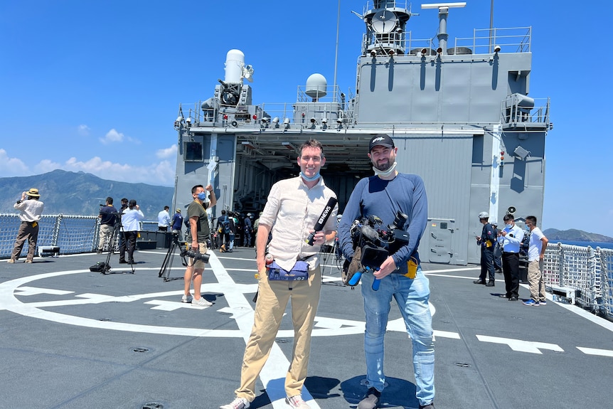 Two men holding ABC camera and microphone standing on deck of navy ship.