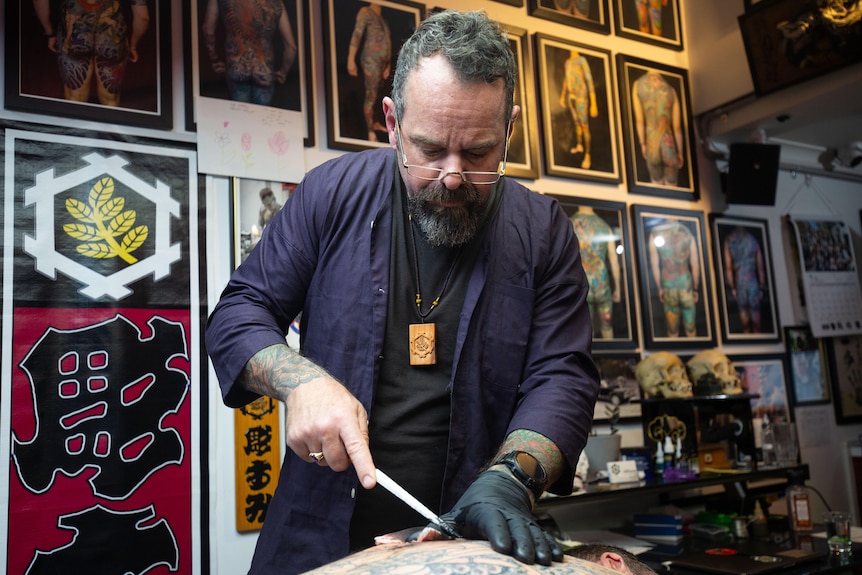 A man in his early fifties is creating a tattoo on the back of another man in his studio using a bamboo stick