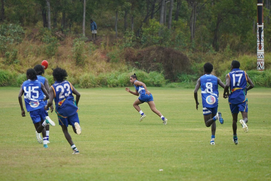 Six players chase after the ball on a green field, with some bush in the background.