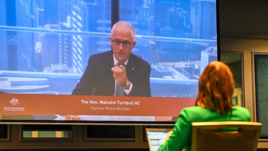 Turnbull speaks video video link while a woman in a green blazer listens. 