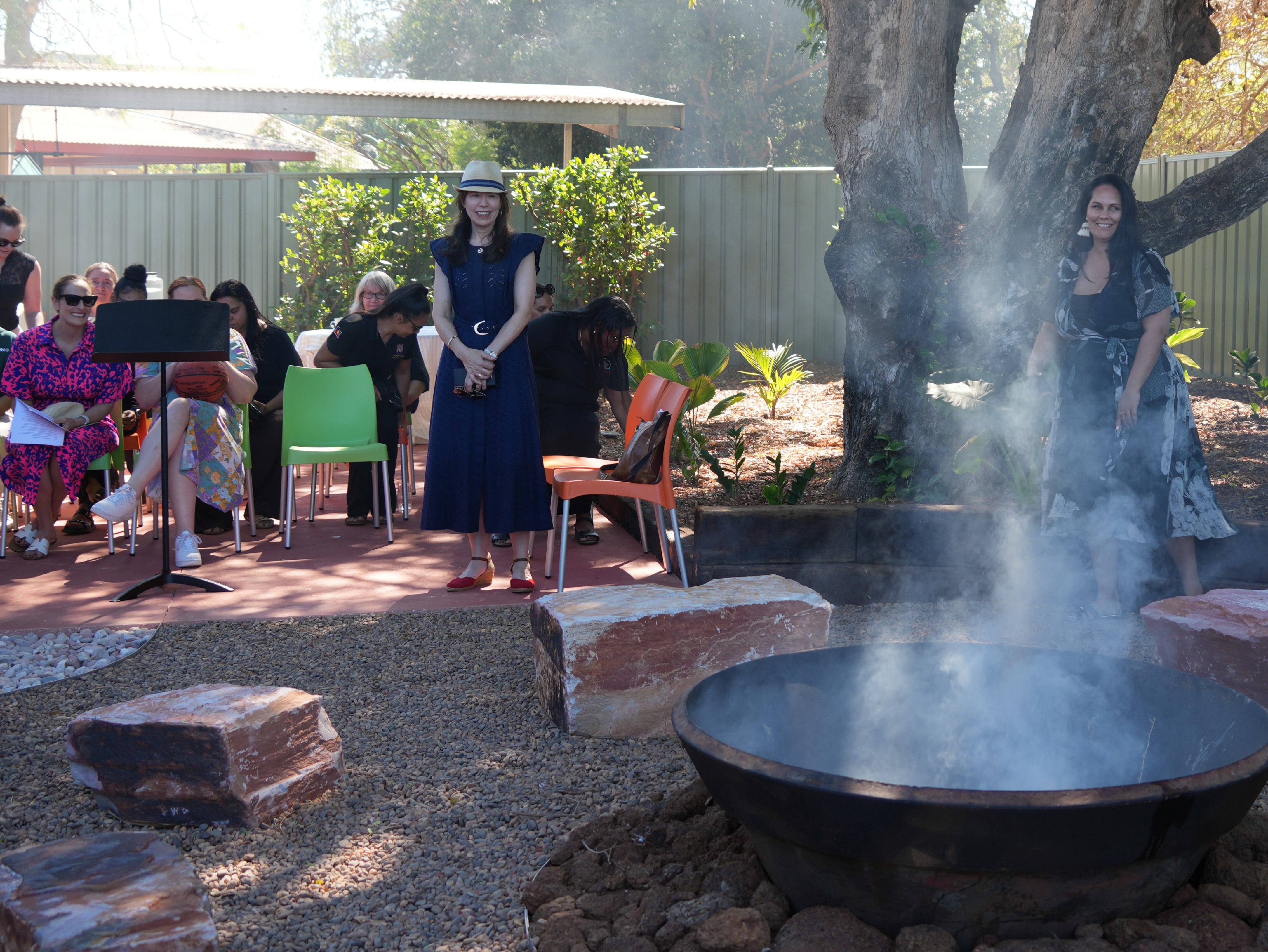 People speak around a smoky campfire under a shady tree