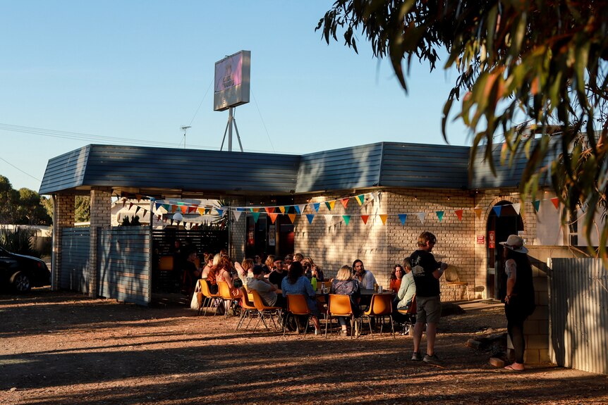 Group of younger people sit outside a motel underneath dappled lighting from nearby tree