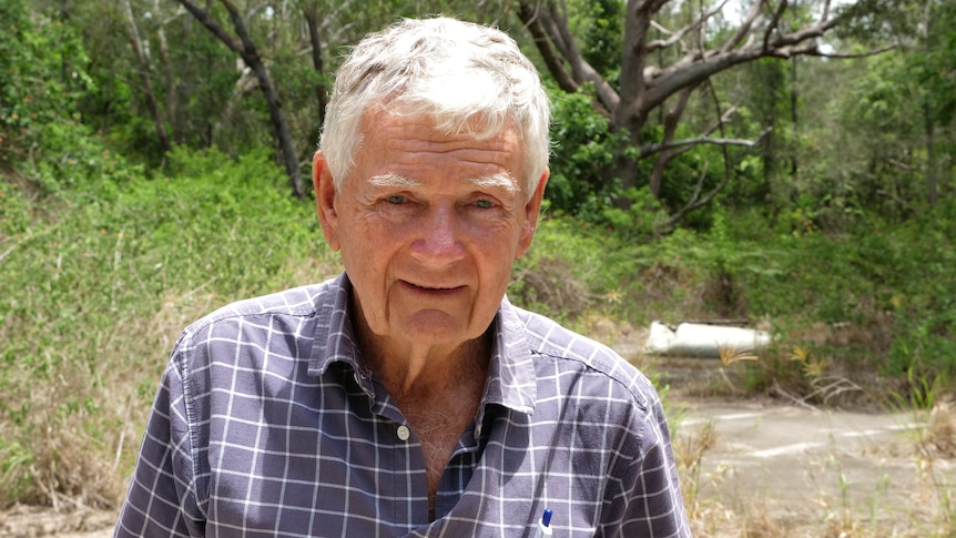 A man in his 70s, short grey hair and a check shirt standing in a clearing the bush.