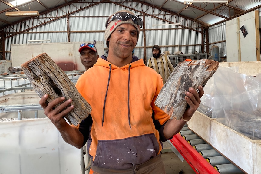 Indigenous man in orange top with beanie holding a log up in each hand in front of two men in background