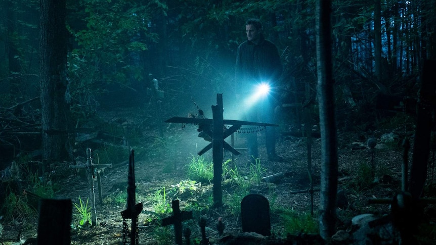 In a dark forest, the actor shines his torch to illuminate a makeshift/DIY graveyard.