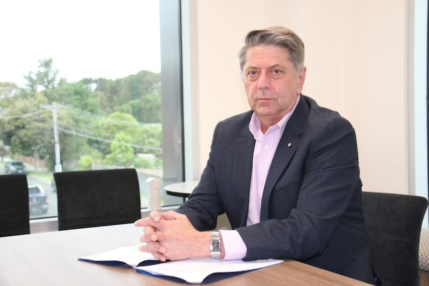 A man in a pink shirt and black jacket sits at a desk in an office.