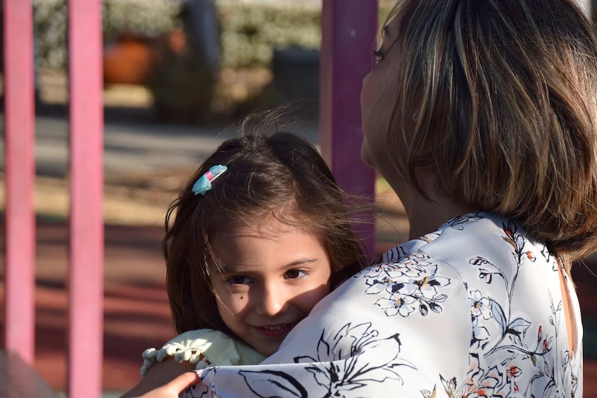 A young girl smiling over the shoulder of her mother as her mother holds her daughter to depict story of life after stillbirth.