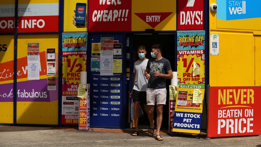 Two men exit a chemist warehouse wearing masks, with signs advertising rapid tests are sold out around the door.