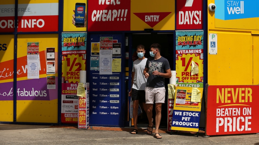 Two men exit a chemist warehouse wearing masks, with signs advertising rapid tests are sold out around the door.