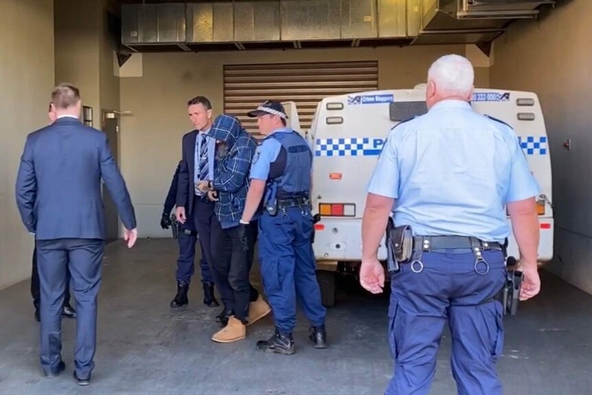 A man with his face covered is escorted from a police wagon into a police station.