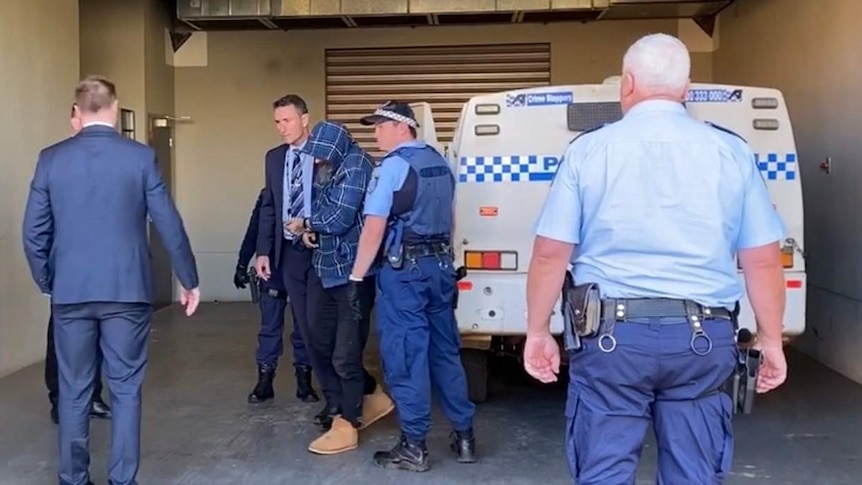 A man with his face covered is escorted from a police wagon into a police station by officers