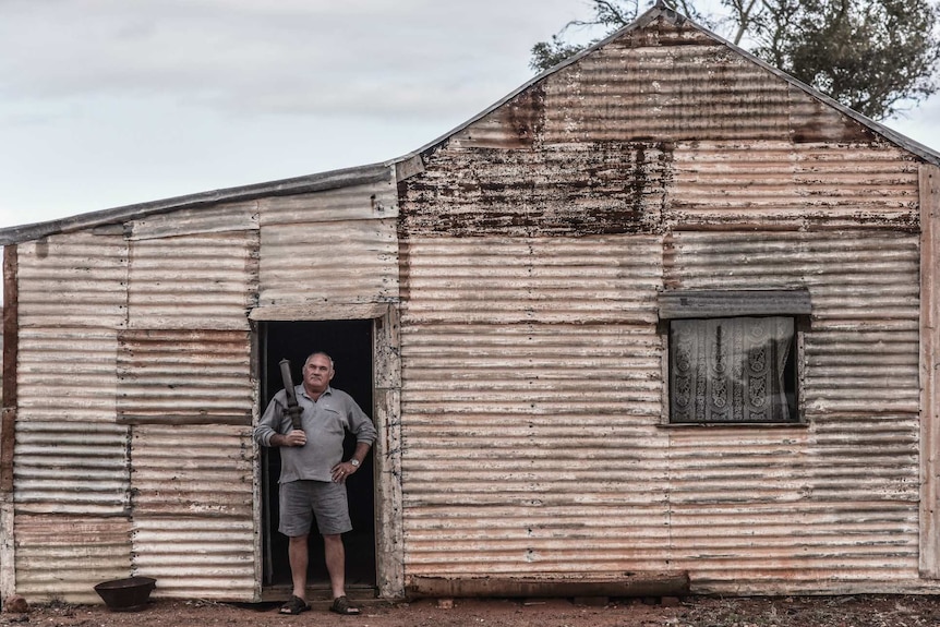 A man in shorts and a grey top stands at the doorway of an abandoned tin shed