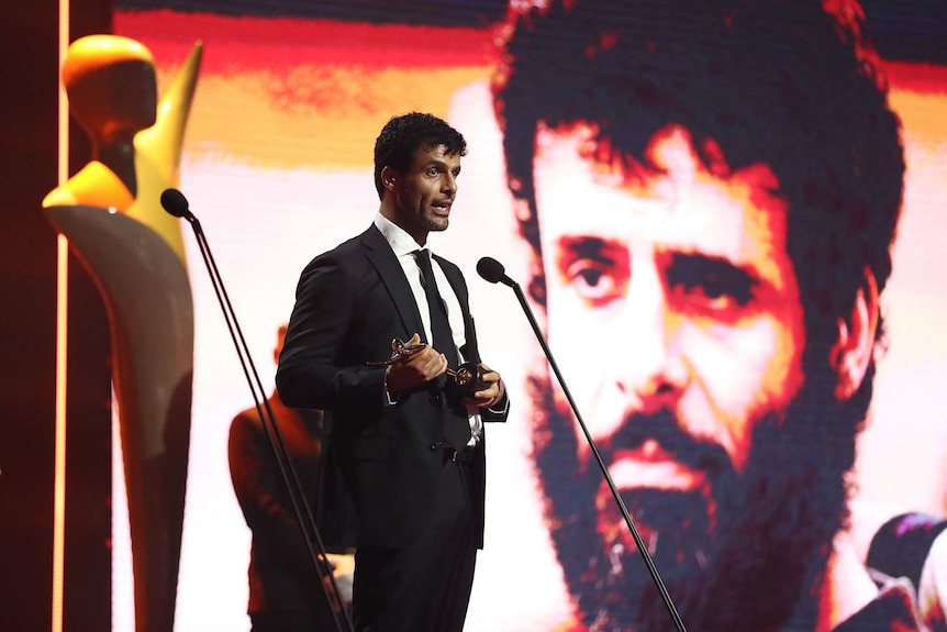 young man on brightly lit stage holding award, speaking, with picture of him in character on large screen in background.