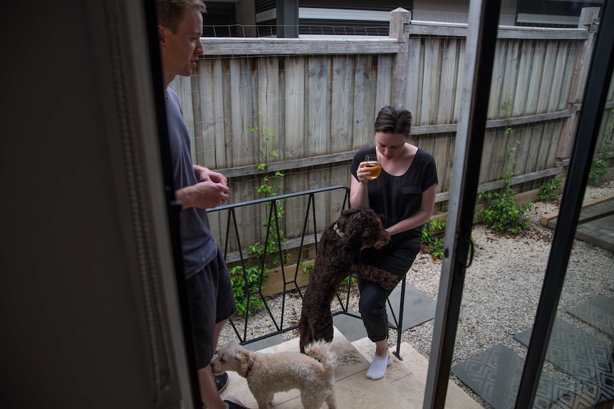Teacher Anita Harding holds a drink in one hand and pats her dog with the other in the backyard, her husband Michael nearby.