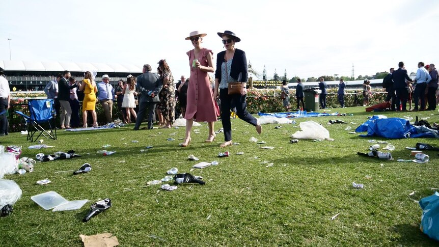 Two women in dresses and hats walk across a green lawn strewn with bags, beer cans and rubbish.