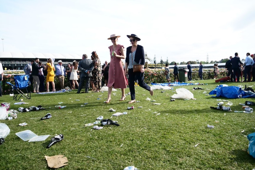 Two women in dresses and hats walk across a green lawn strewn with bags, beer cans and rubbish.