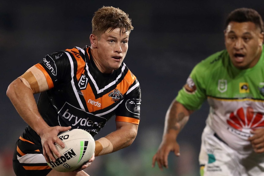 A man in a Tigers jersey runs with the ball on a rugby league field.