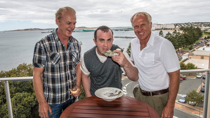 Three men on hotel balcony high up, middle one eating a very large oyster, county town in background.