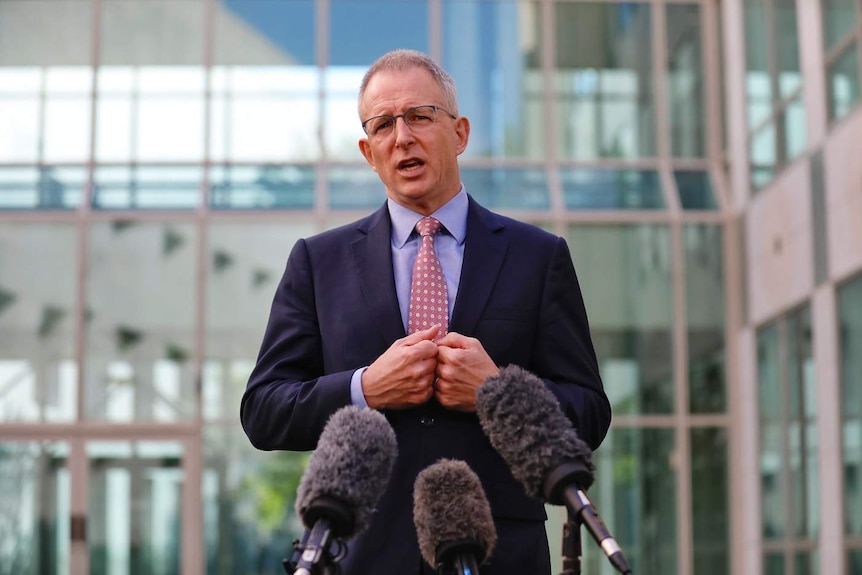 Paul Fletcher speaks in front of a bank of microphones.