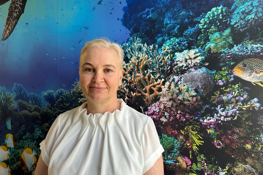 A woman with blonde hair and dark eyebrows standing infront of a large photo of coral. 