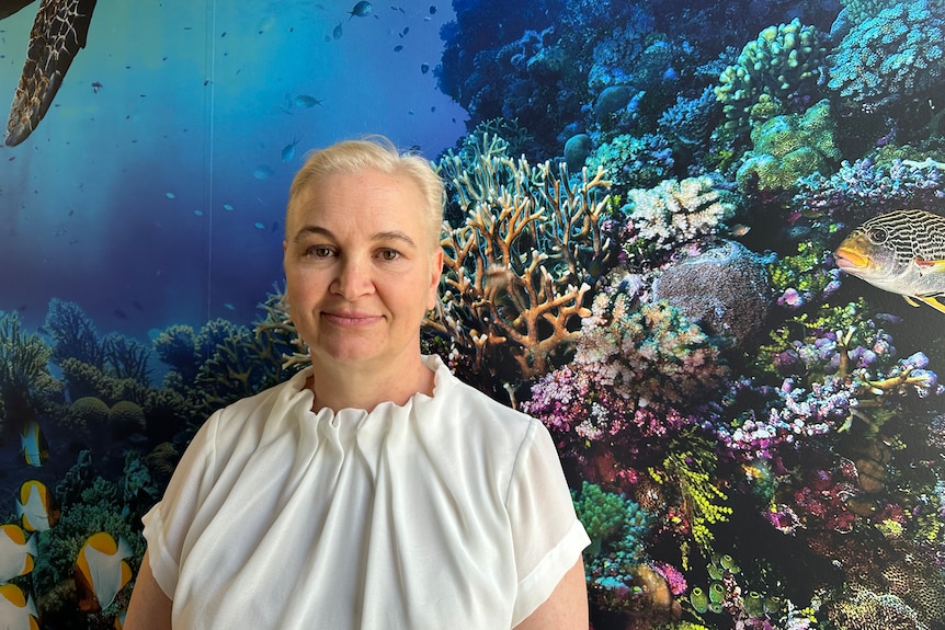 A woman with blonde hair and dark eyebrows standing infront of a large photo of coral. 