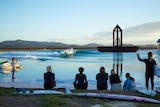 Six people with surfboards and wetsuits look at over a lake with waves and a massive metal structure in the centre.