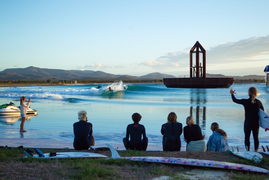 Six people with surfboards and wetsuits look at over a lake with waves and a massive metal structure in the centre.