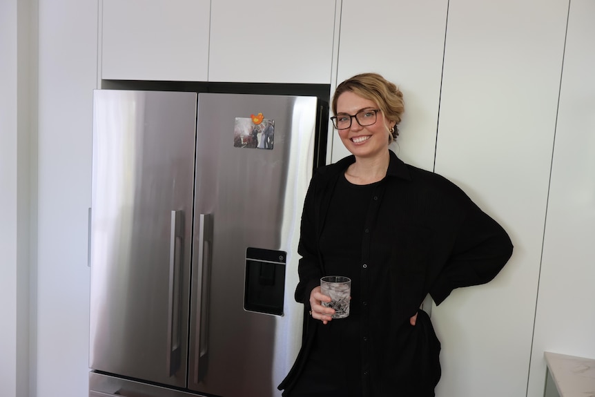 Amy Thunig stands next to a large silver fridge with a glass of water in her hand.