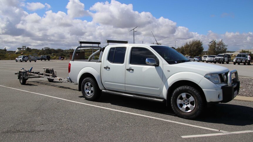 A white 4WD utility sits parked in a car park with a boat trailer connected to the back of it.