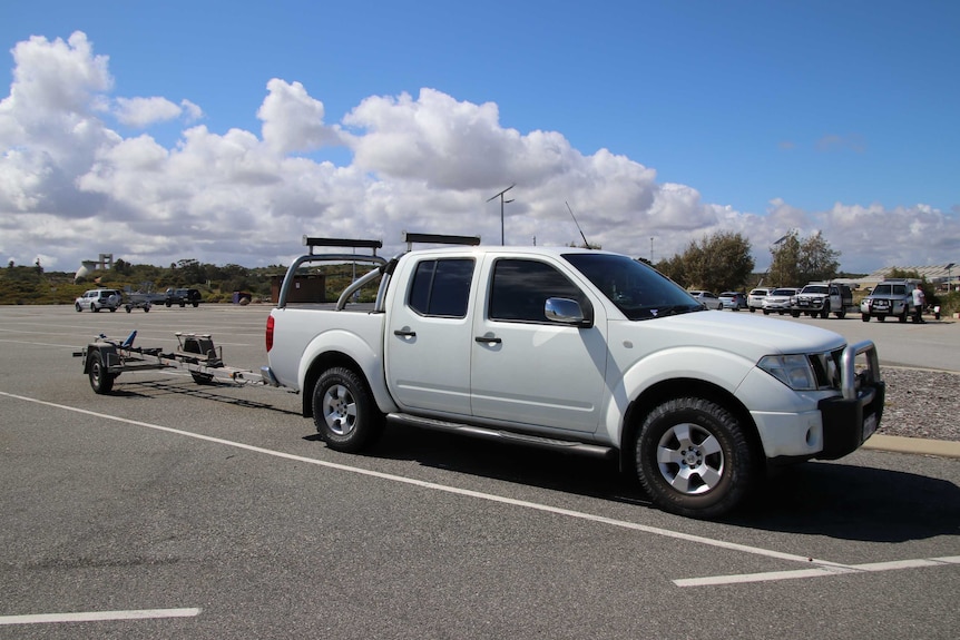 A white 4WD utility sits parked in a car park with a boat trailer connected to the back of it.