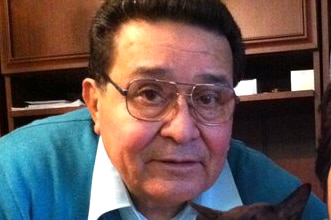 An older man in glasses and a blue jumper poses for a photo, with a black cat in the foreground.