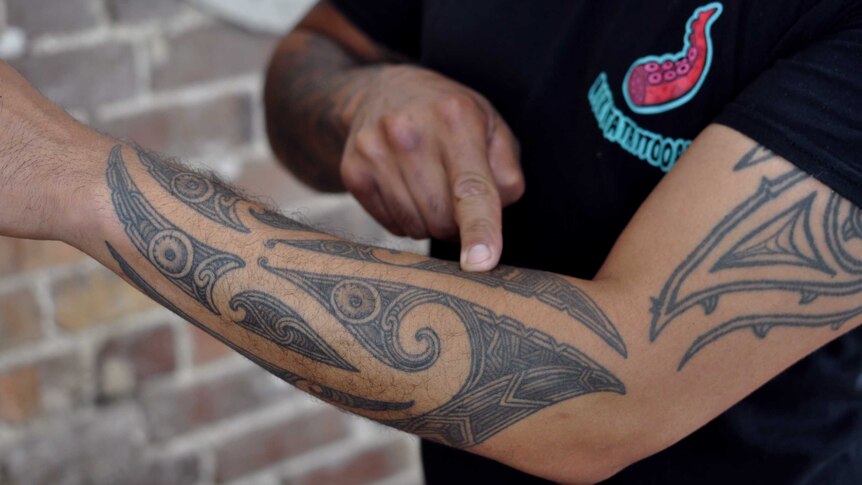 A Maori man whose face is not in shot points at a traditional tattoo on his arm, explaining its meaning.