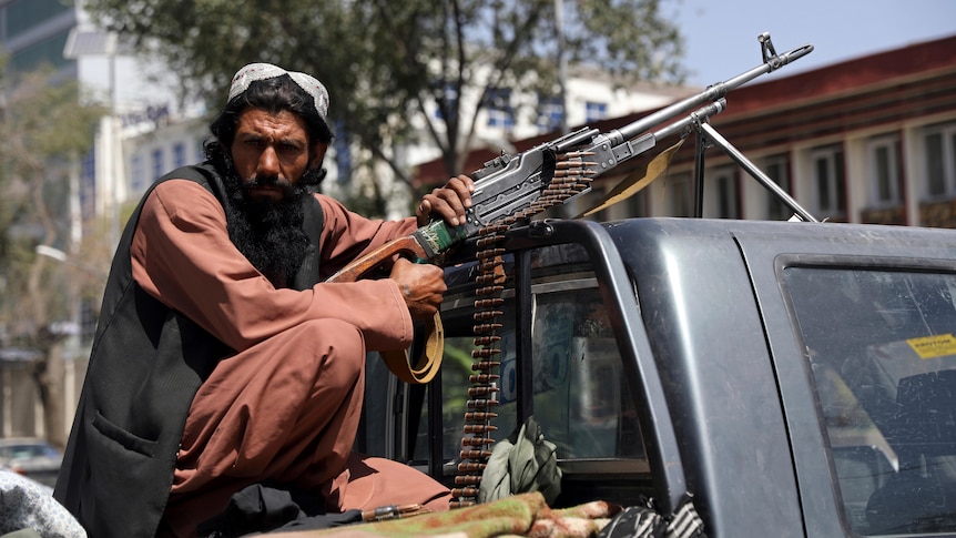 A bearded man with a giant rifle sits in the tray of a ute, looking at the camera 
