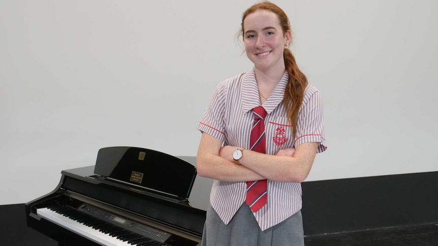 A teenage girl in school uniform standing next to a black piano on a school stage