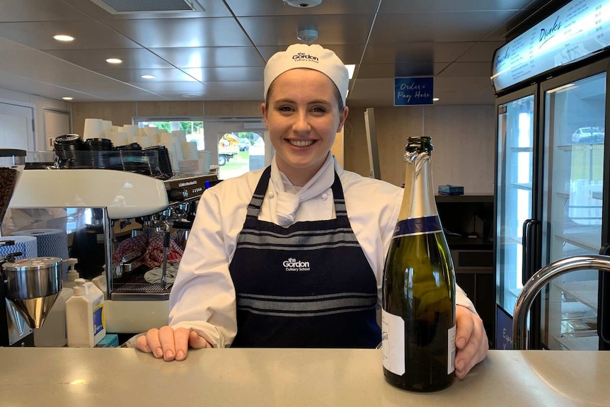 A woman wearing a white chef uniform and blue apron serves champagne in the ferry cafe.