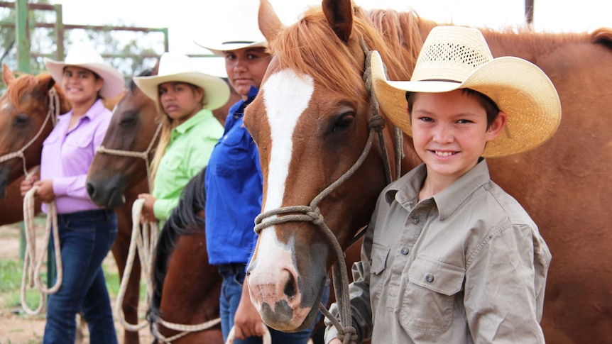 A line of smiling children wearing hats, each standing next to a horse.