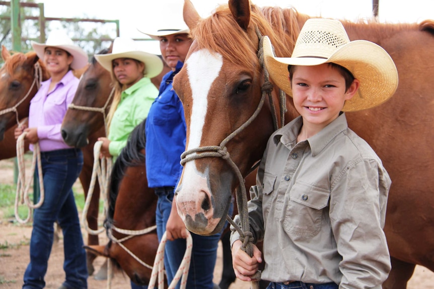 A line of smiling children wearing hats, each standing next to a horse.