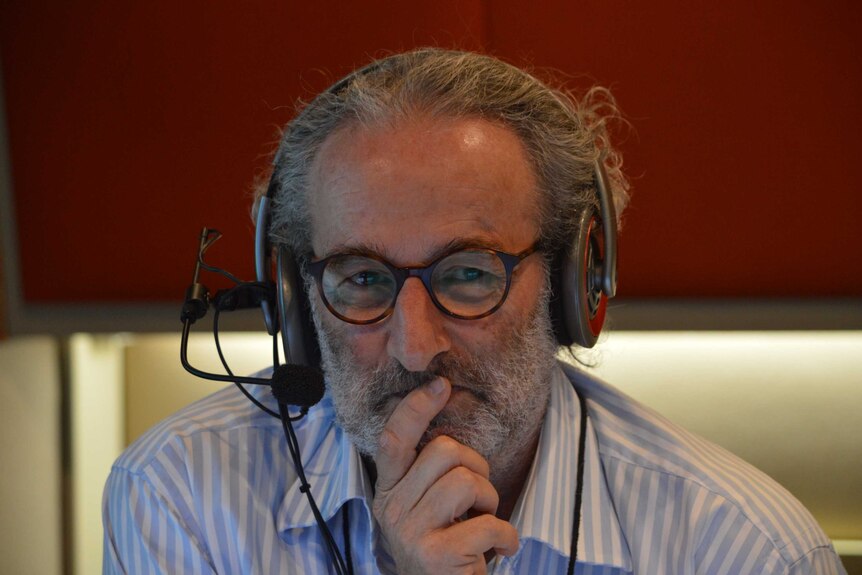 Tight shot of Jon Faine with headphones on in studio and finger on mouth as though thinking.