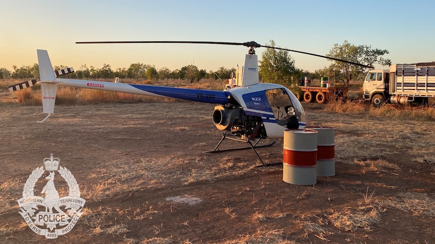 a helicopter in a paddock with drums and trucks nearby.