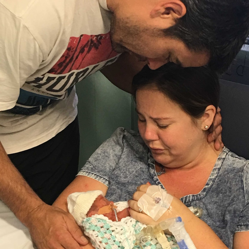 A mother cries over her baby in a hospital room while a man embraces them.
