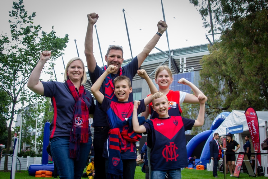 A mum, dad, and three kids in Melbourne football club jumpers stand cheering outside the MCG