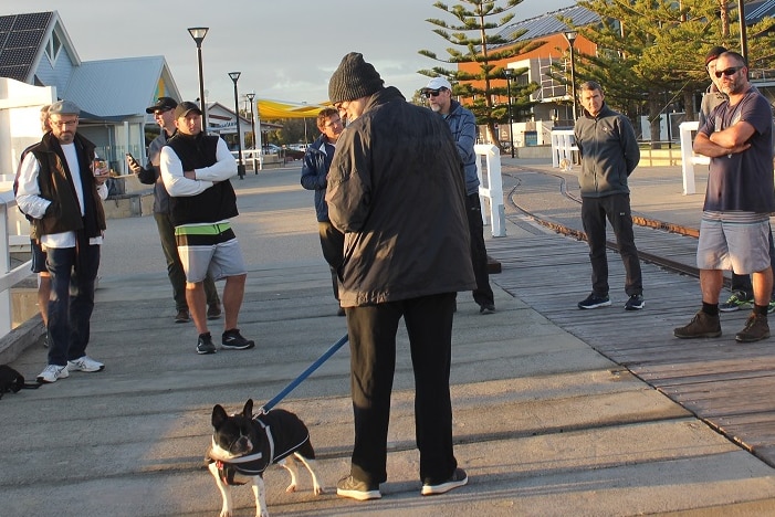 A group of men standing talking on the Busselton jetty, with dogs. It's cold and they have hats and jackets on