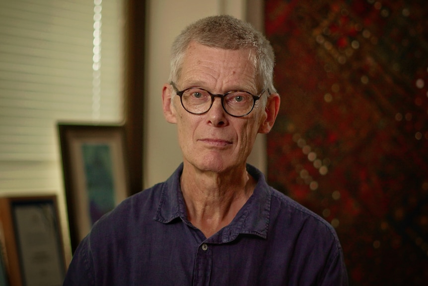 Jeremy Hayllar wearing a linen blue shirt standing in front of a window with frames on the sill and maroon fabric behind him