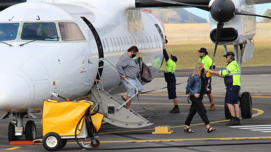 Two passengers disembark from a small airplane, lots of people in hi-vis vests stand around them