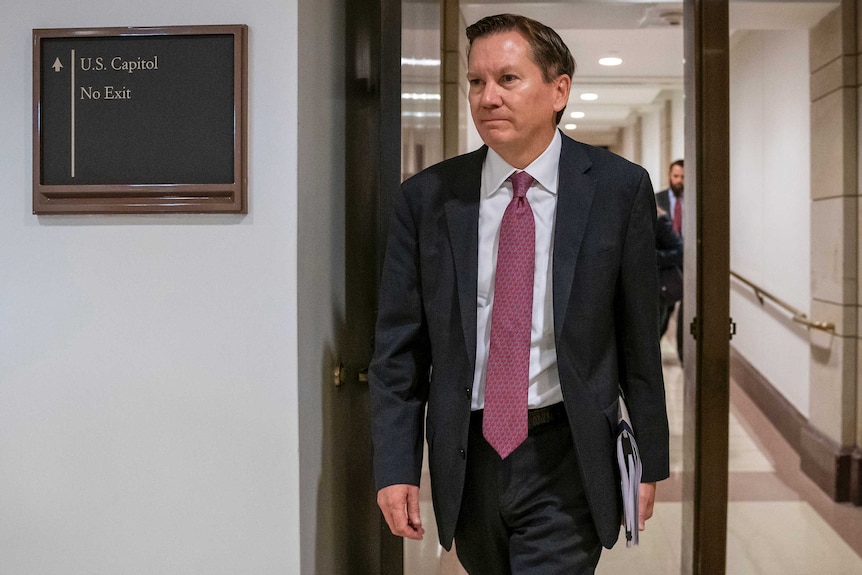 Michael Atkinson, wearing a suit and carrying a folder, walks into a room.