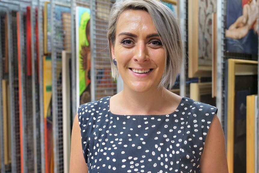 A smiling woman with  short blonde hair, brown eyes, stands in an art store room with racks behind.