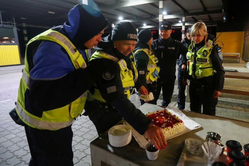 People in high-vis vests and police uniforms stand around a table with cake and coffee cups