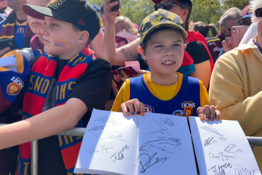 A boy wearing a Lions guernsey and a yellow and black Pokemon cap holds two pages of autographs up while surrounded by fans.